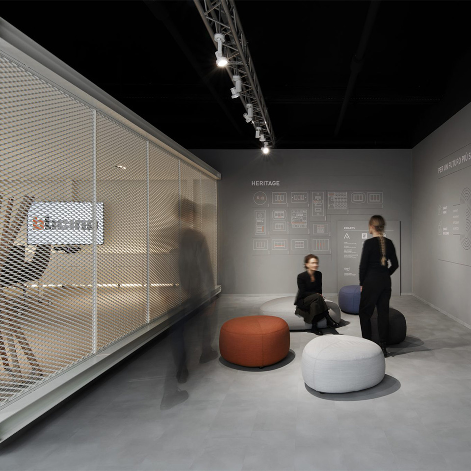 Modern showroom at Bticino headquarter with mesh wall, seating ottomans, and blurred figures in a minimal grey space.