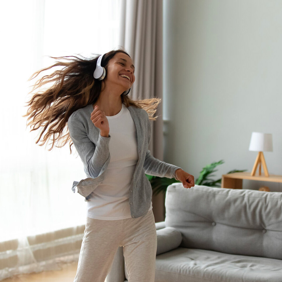 Woman wearing headphones dances and smiles in a living room with natural light, sofa, and table lamp in the background.