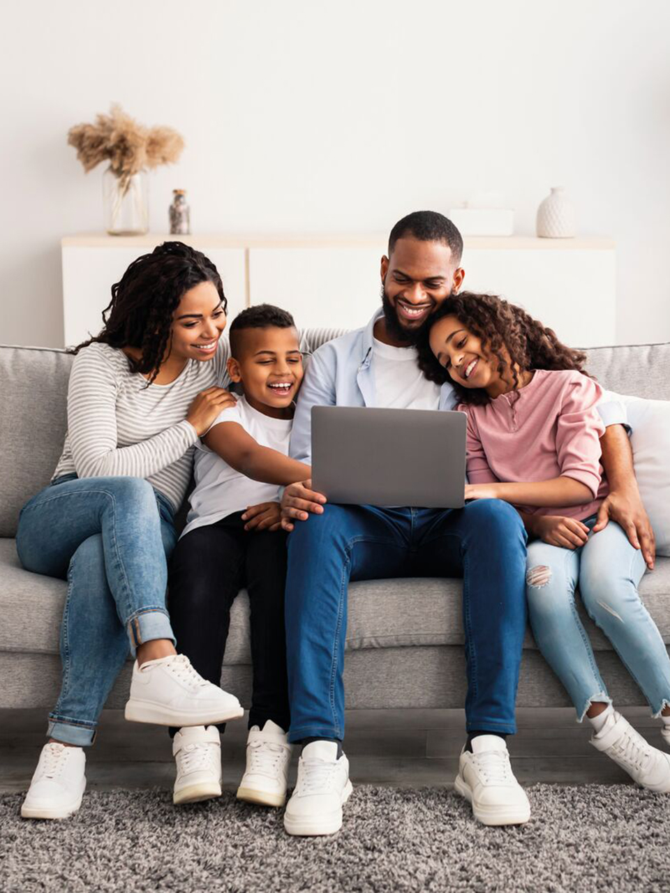 A family of four sits on a couch, smiling and looking at a laptop together in a living room.