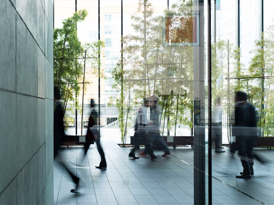 People walk through a modern glass-walled lobby with indoor trees and blurred motion indicating movement.