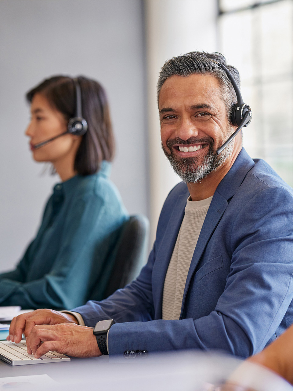Man in a blue suit with a headset smiling at desk; woman with headset working in background.