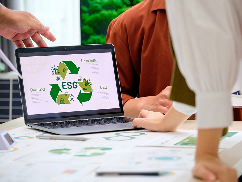 A laptop screen displays an ESG infographic as people discuss documents around a table.