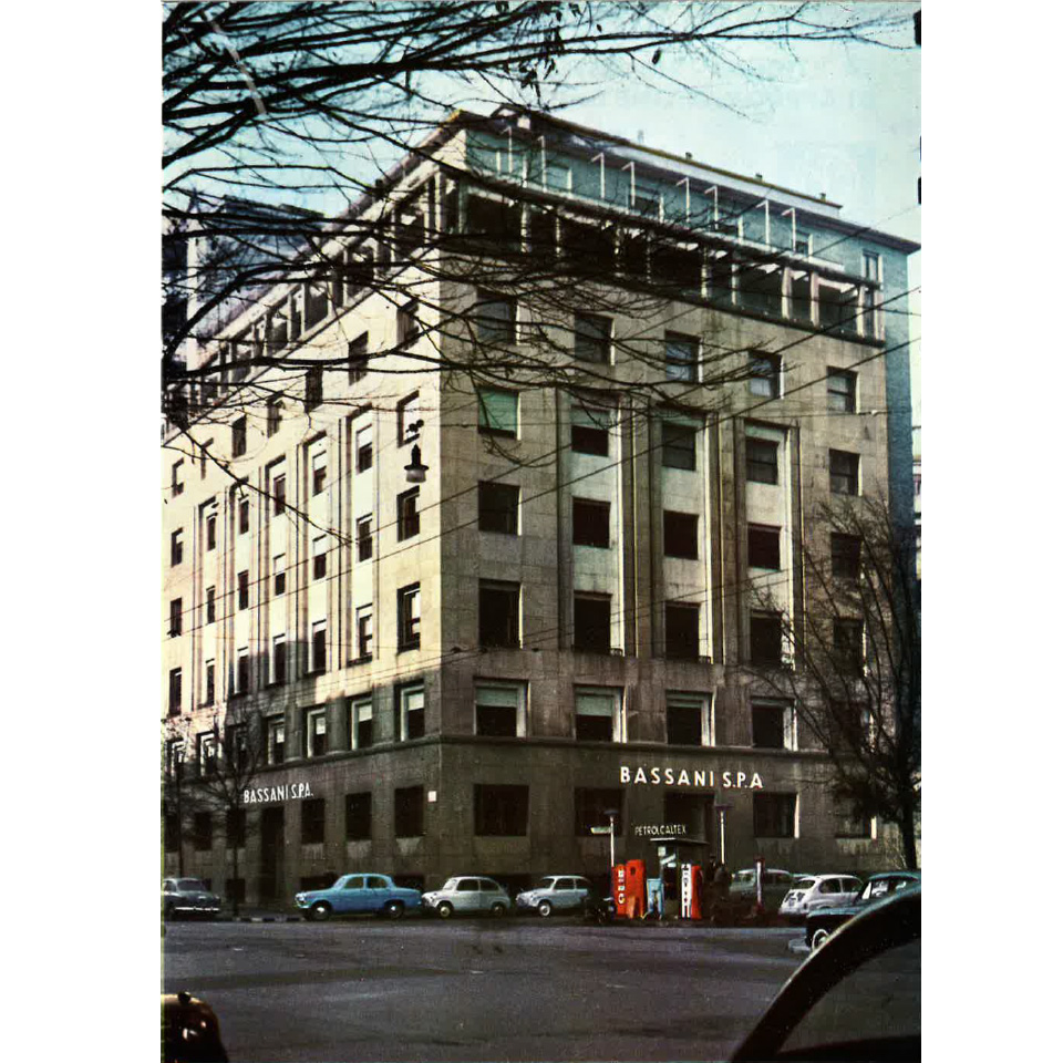 A 60s multi-storey office building in Milano with BASSANI S.P.A. signage, parked cars and trees.
