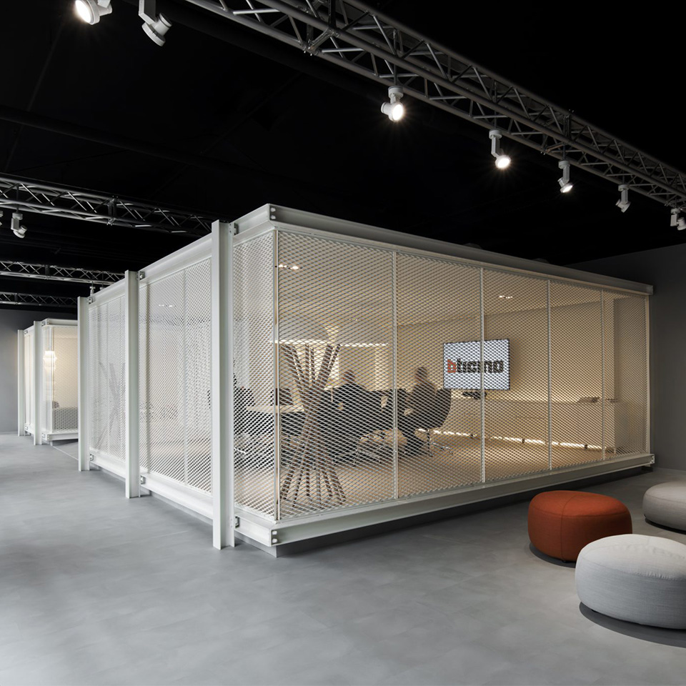 Modern office space with mesh-walled meeting room, lounge chairs, a TV screen, and round cushioned stools.