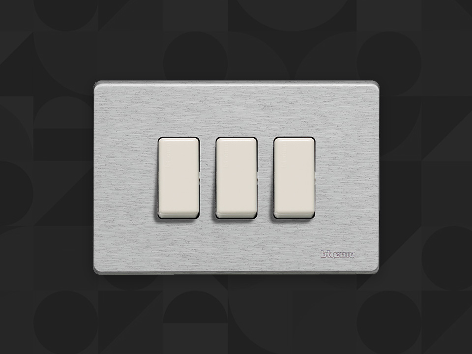 BTicino Magic silver plate with 3 white switches, on a dark patterned background.