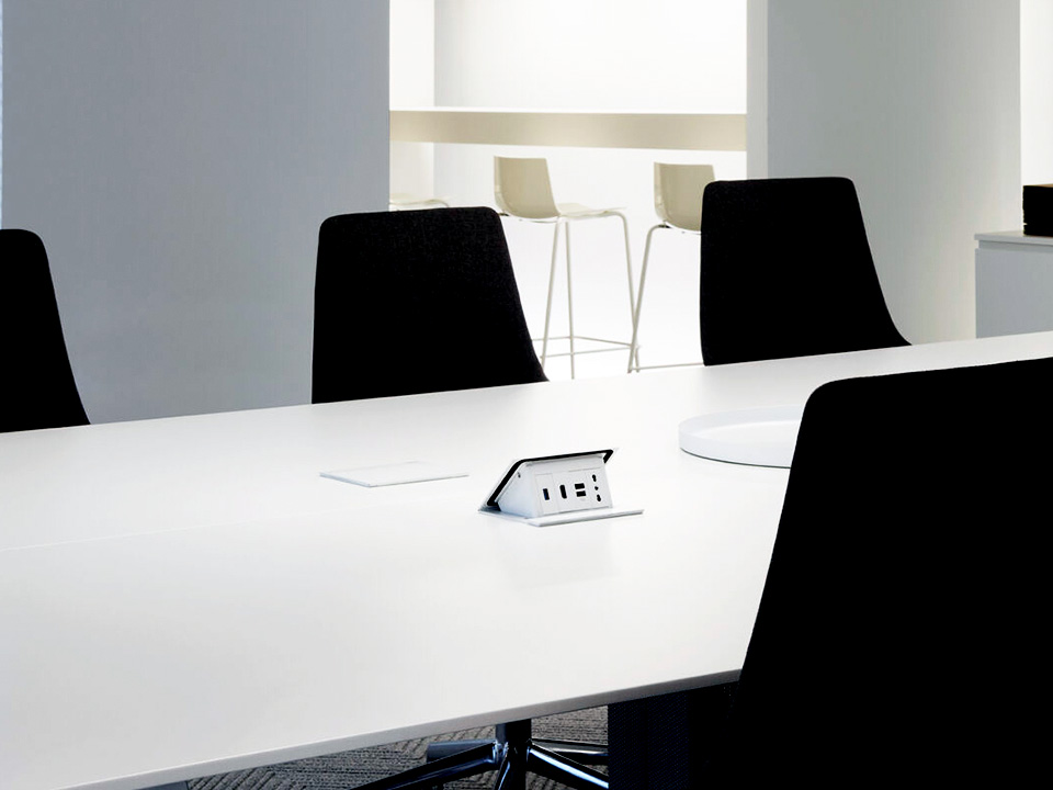 A modern conference room with a white table, black chairs, and power outlets on the table; stools in the background.