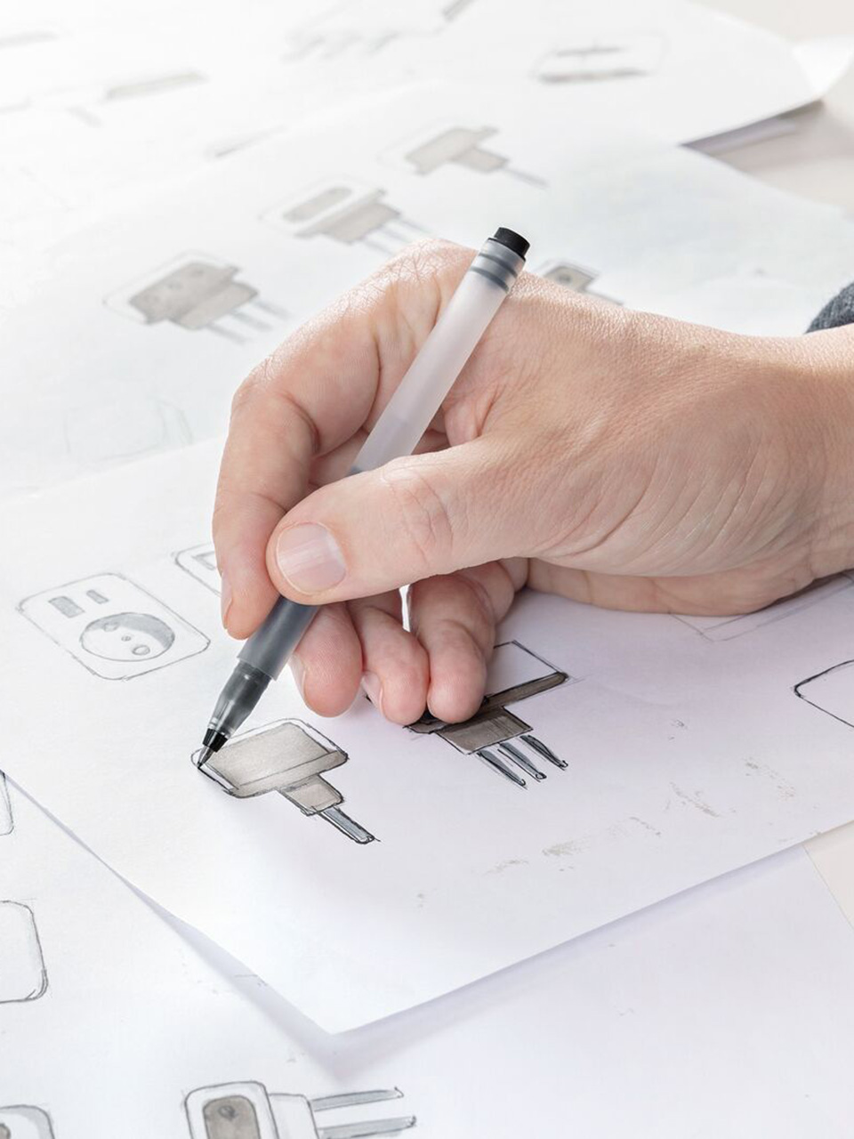 A hand draws sketches of electrical plugs on white paper with a black pen.