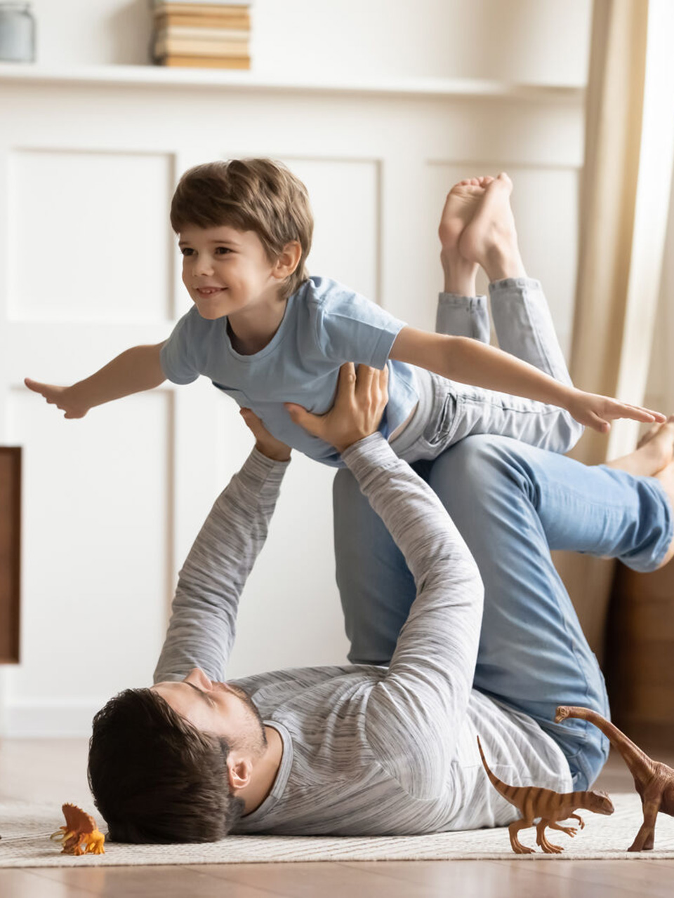 Adult lying on the floor lifts a smiling child in the air as toy dinosaurs are scattered around them in a living room.