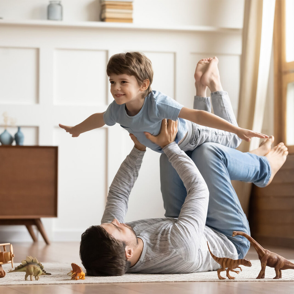 Adult lying on the floor lifts a smiling child in the air as toy dinosaurs are scattered around them in a living room.