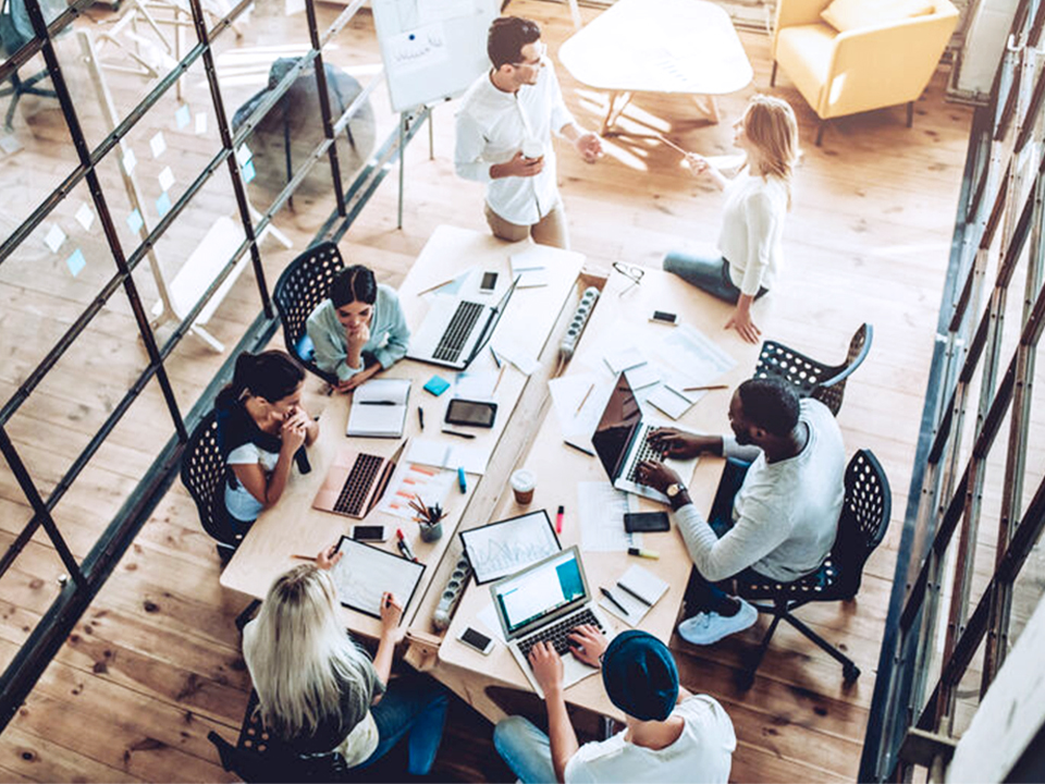 A group of people working together at a table with laptops, notebooks, and papers in a modern office space.