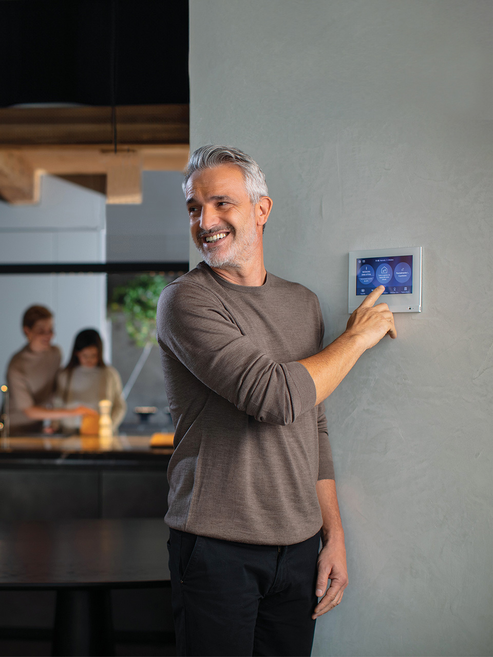 A man adjusts the BTicino MyHome home automation system screen, while two people talk in the kitchen background.