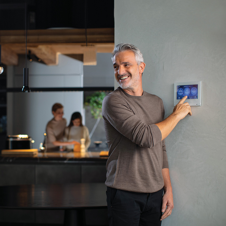 A man adjusts the BTicino MyHome home automation system screen, while two people talk in the kitchen background.