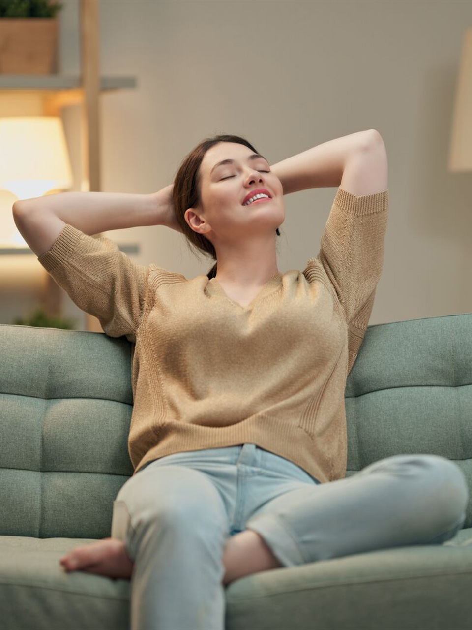 Woman sitting on a sofa with her hands behind her head, eyes closed, appearing relaxed and content.
