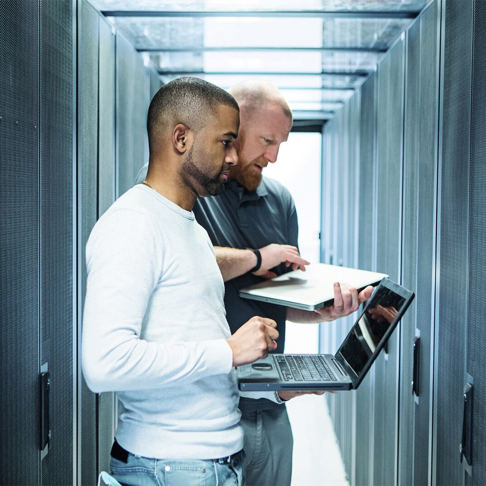 Two men standing in a server room, each working on a laptop among tall server racks.
