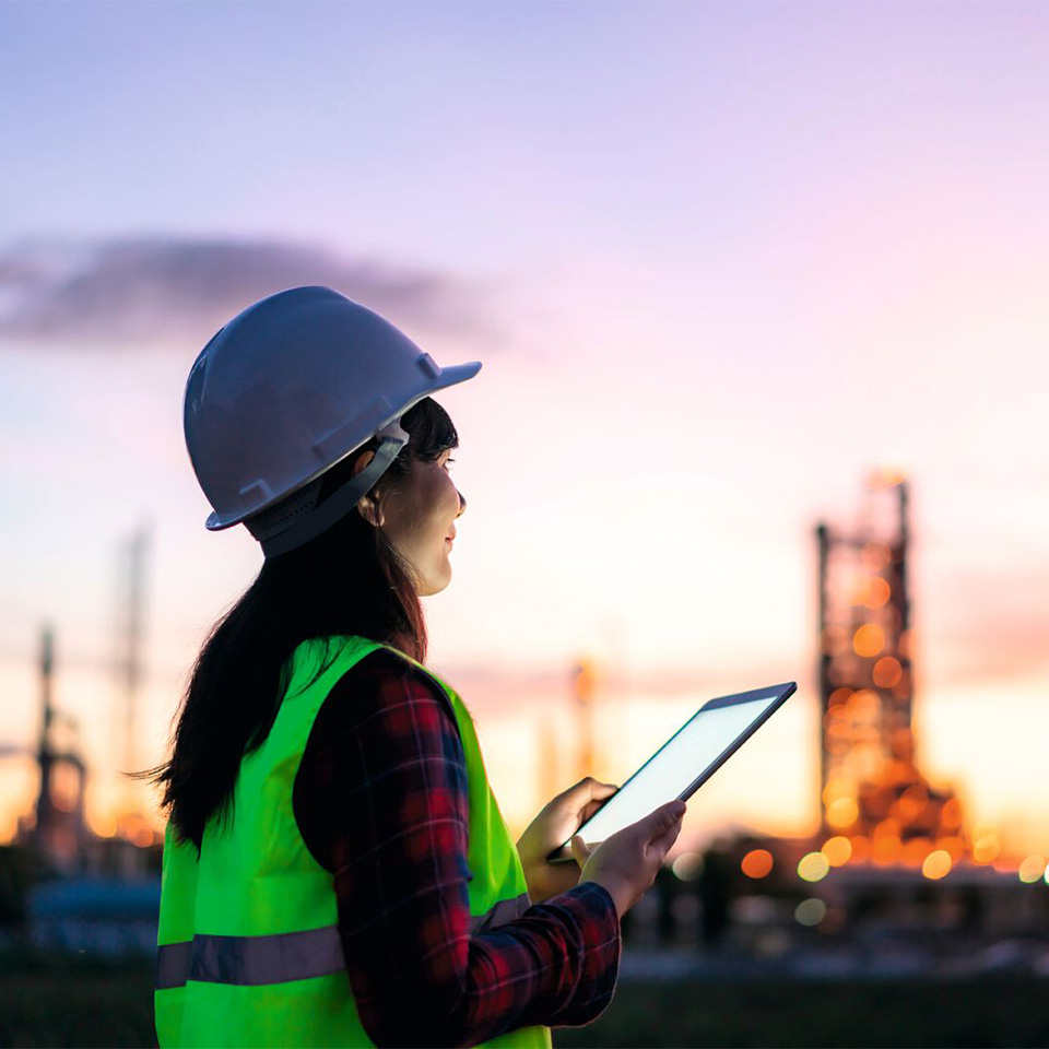 A person in a hard hat and safety vest uses a tablet at an industrial site during sunset.