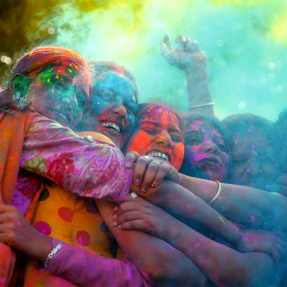 A group of people celebrate Holi, smiling and hugging, covered in colorful powder with vibrant clouds around them.