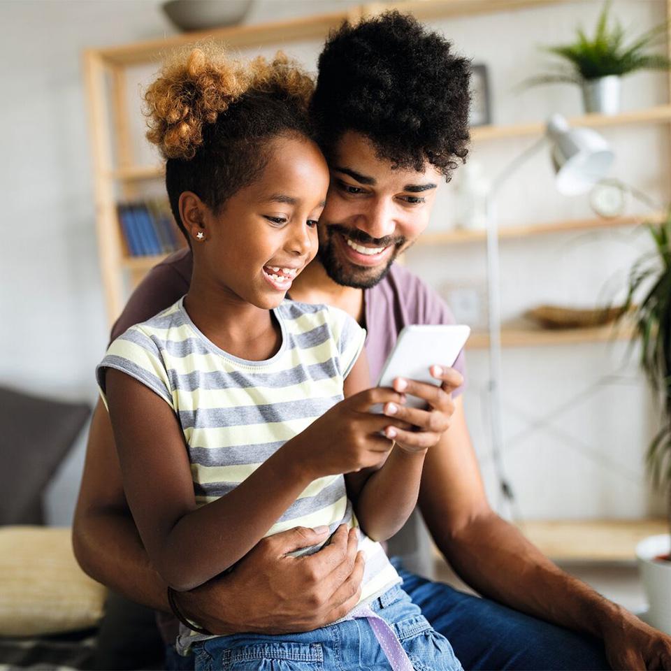 A man and a young girl smile while looking at a smartphone together in a living room.