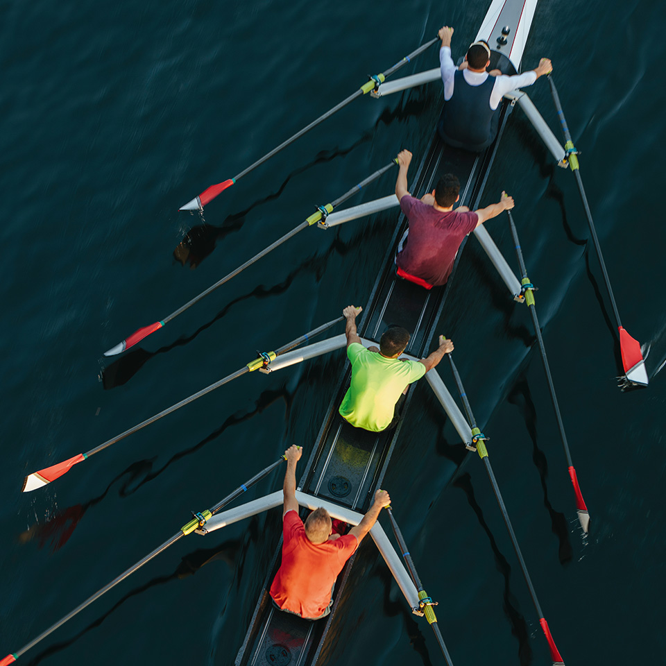 Four rowers paddle in sync on a racing shell in calm, dark water, viewed from above.