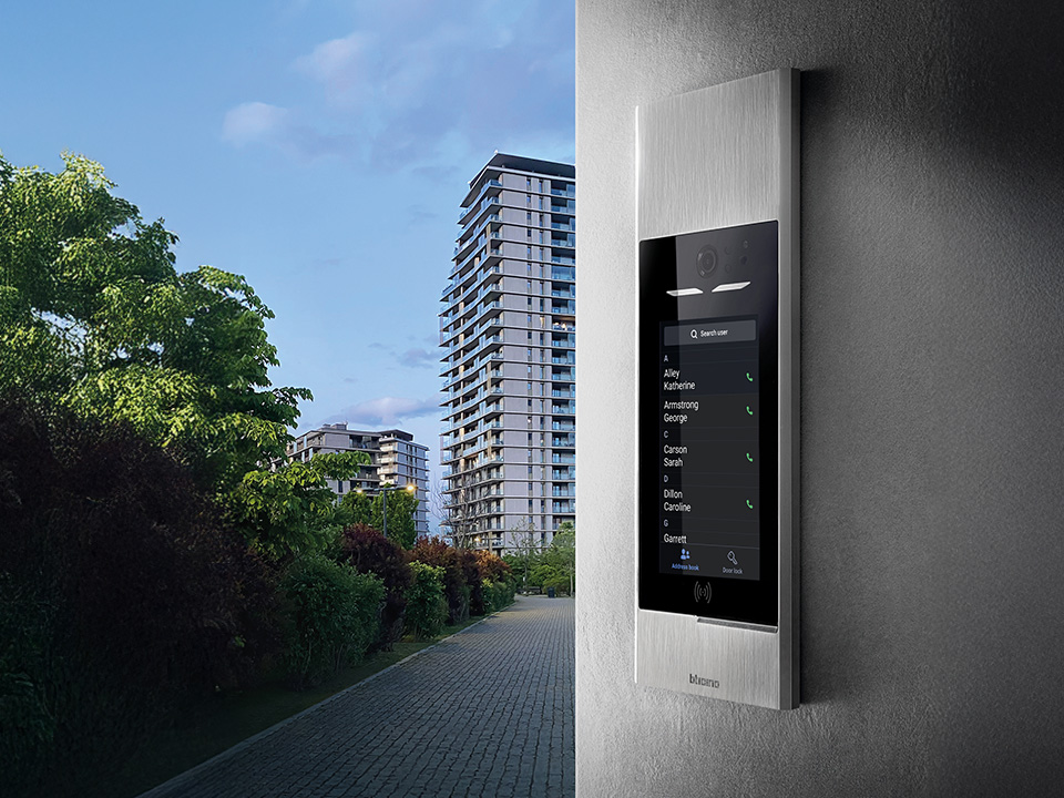 Wall-mounted Linea 5000 video intercom beside a walkway lined with trees and modern apartment buildings.