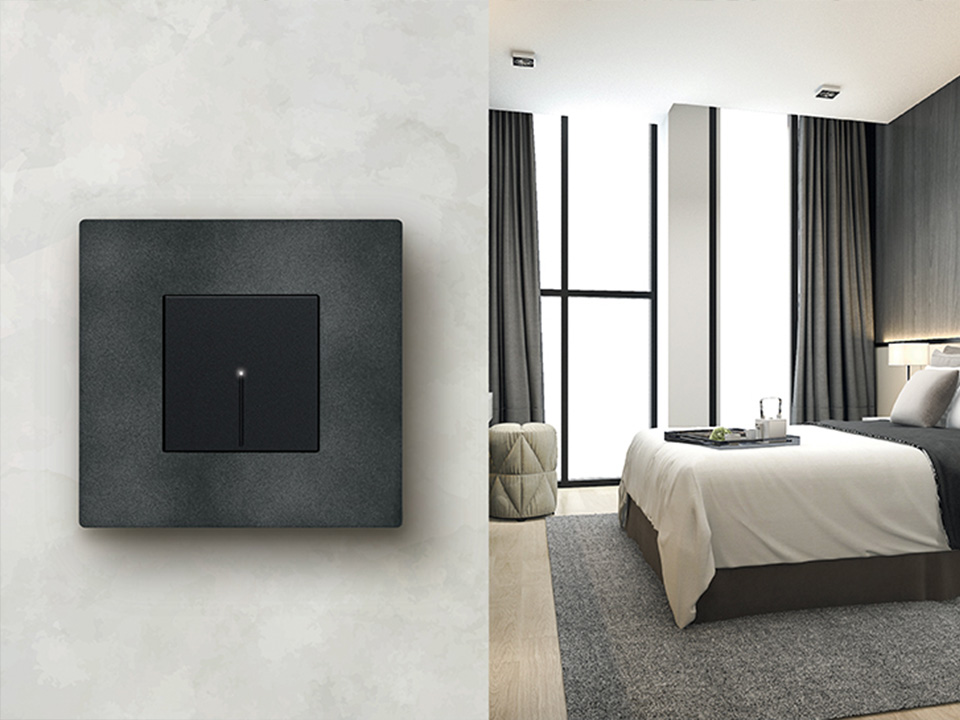 BTicino Light Now 2-module light switch in dark grey, installed on a textured wall beside a bedroom.