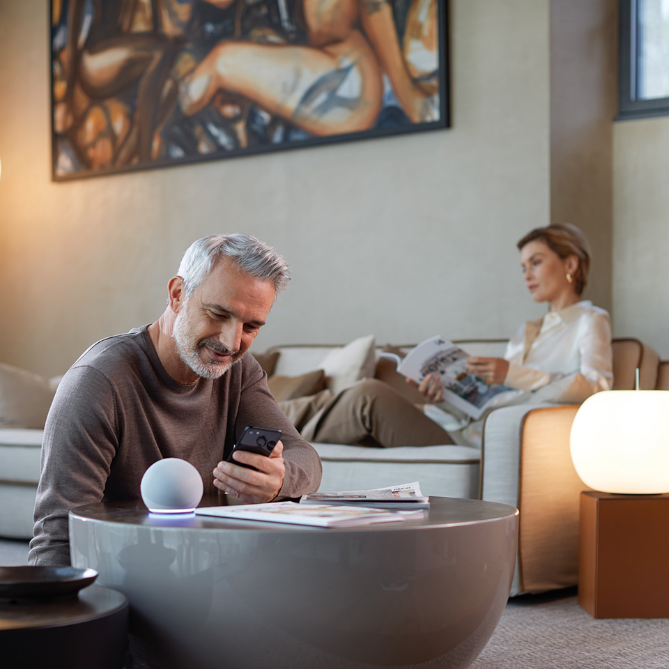 A man uses his phone at a coffee table with a smart speaker; a woman reads on a sofa in the background.