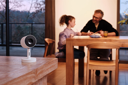 Close-up image of an Advance smart camera in white. In the background, a father and daughter are sitting at a table.