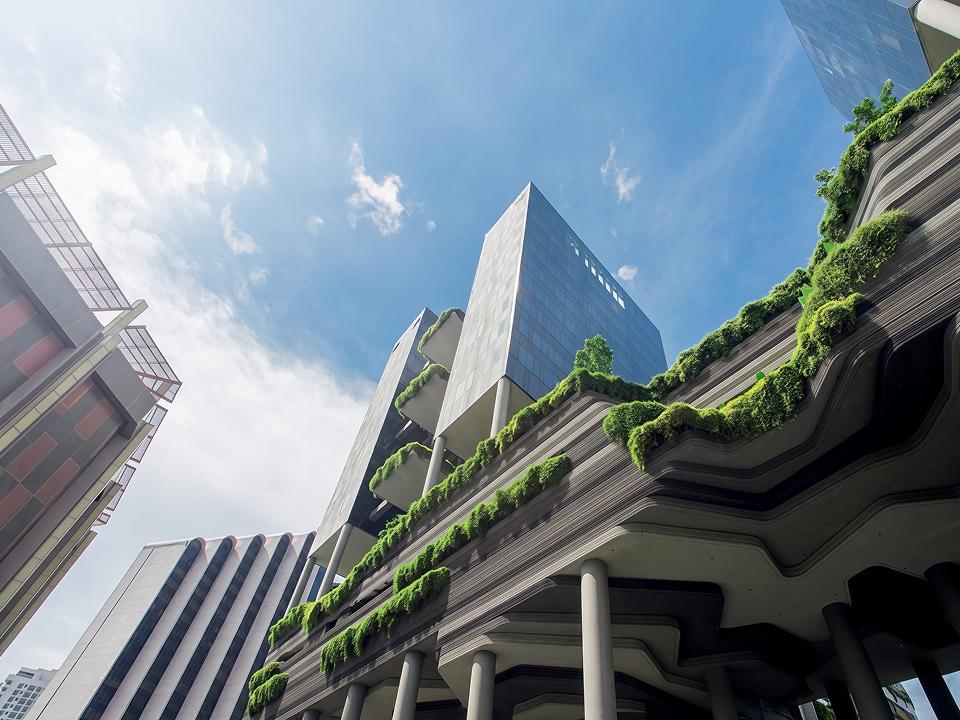 Modern buildings with greenery cascading from terraces under a blue sky, viewed from a low angle.